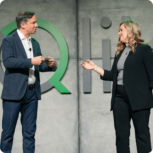 A man in a blue blazer and a woman in a black blazer are smiling and interacting on stage in front of a large grey and green Qlik logo. The man is holding a microphone and gesturing towards the woman.