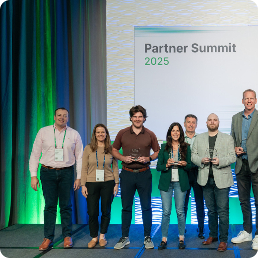 A group of seven people smiling for a photo on a stage in front of a screen displaying "Partner Summit 2025." Three people in the center are holding clear, circular glass awards.