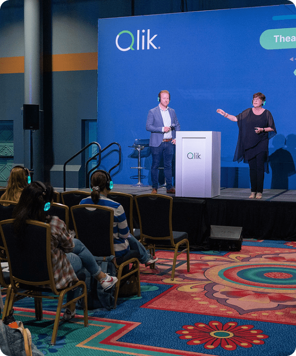 Two presenters speaking on a Qlik stage to a small, seated audience wearing headsets.