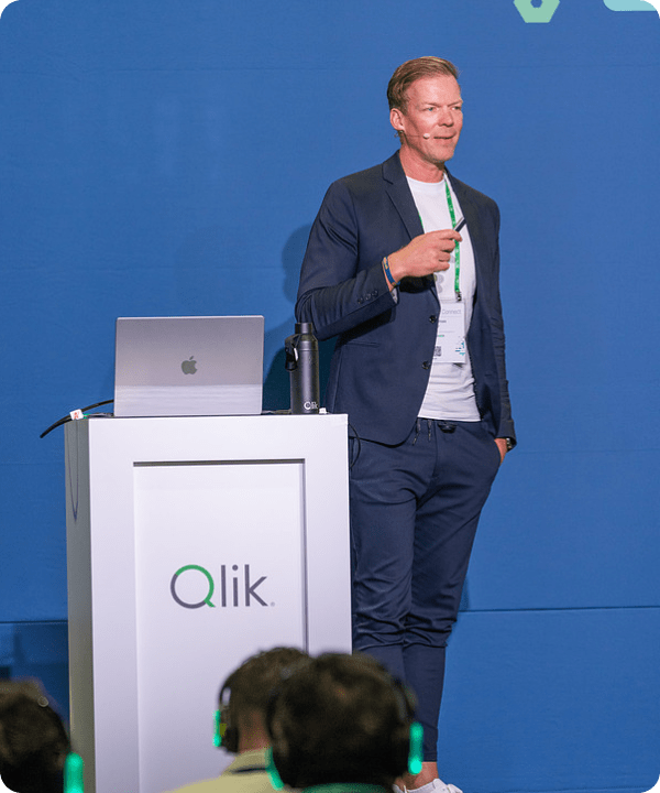A male presenter in a suit and white t-shirt stands at a white Qlik podium with a laptop, speaking to an audience at a conference. The audience members in the foreground are wearing headphones.