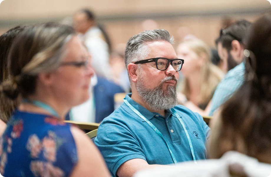 A man with a grey beard and glasses wearing a conference lanyard sits attentively in an audience.