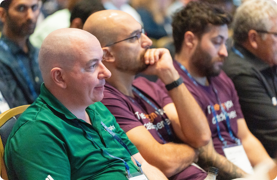 Two men listening attentively in a conference audience, one in a green polo and another behind him in a purple t-shirt.