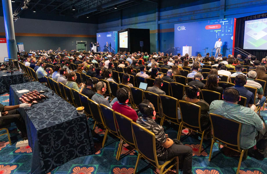 A large audience attends a Qlik Connect presentation in Theater 2, listening to a speaker on stage while wearing headsets for an immersive data analytics session.