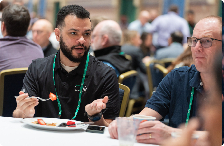 Two men having a conversation at a round table during a conference meal, with one wearing a Qlik polo.
