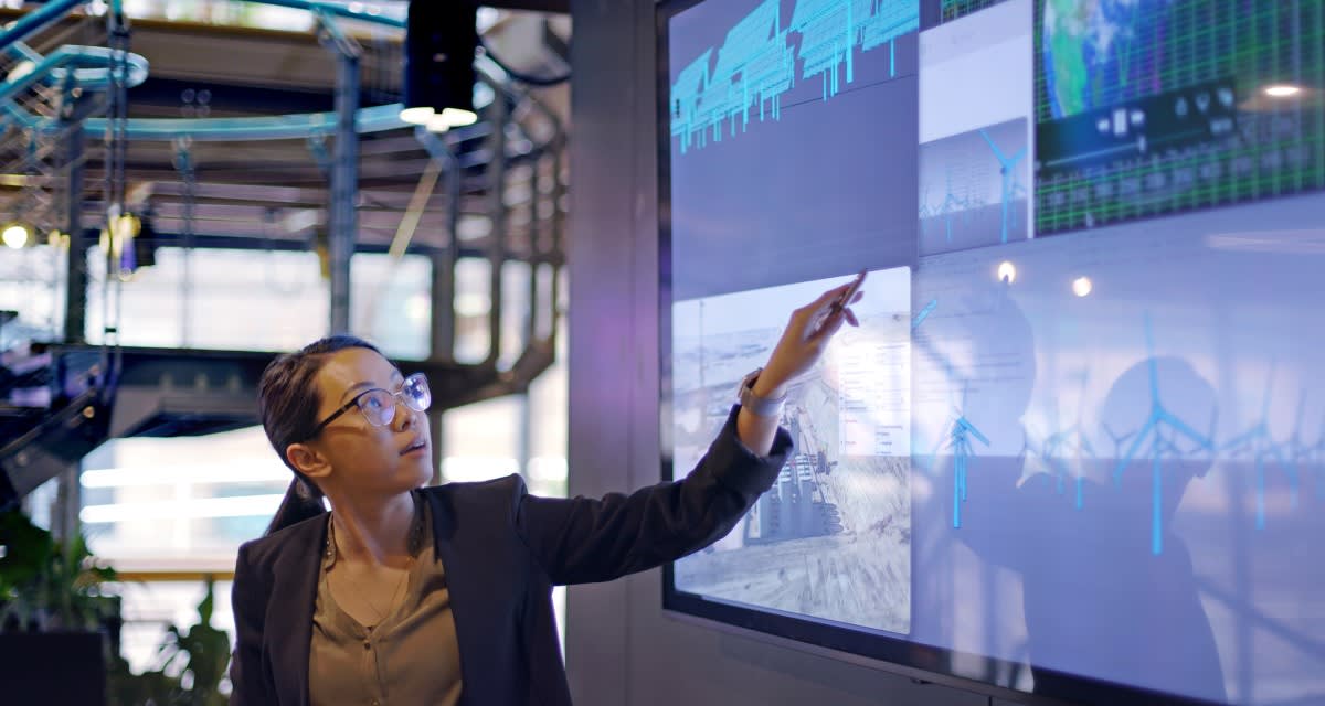 A person in a business suit points at a large touch screen displaying multiple data charts and graphs in a modern office setting.