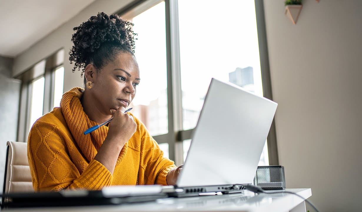 A woman in a yellow sweater sits at a desk, focused on her laptop while holding a pen in her hand.