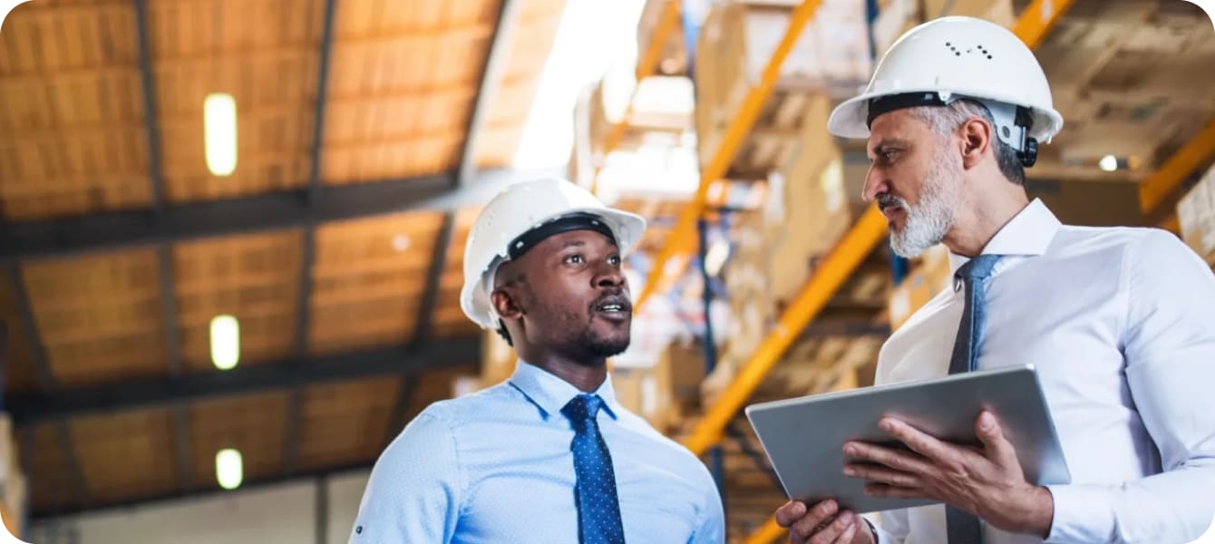 Image of two men in hard hats engaged in conversation inside a warehouse, surrounded by shelves and industrial equipment.