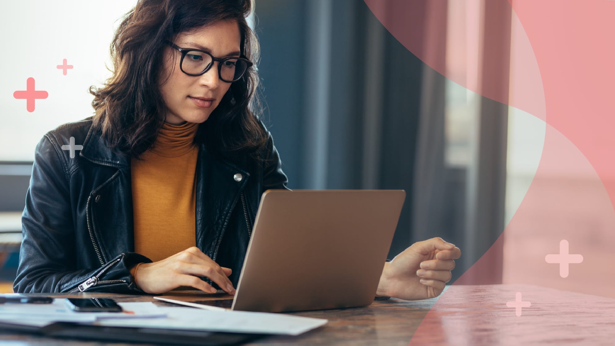 A woman wearing glasses and a black jacket works on a laptop at a table with papers and a pen, against a background with pink accents.