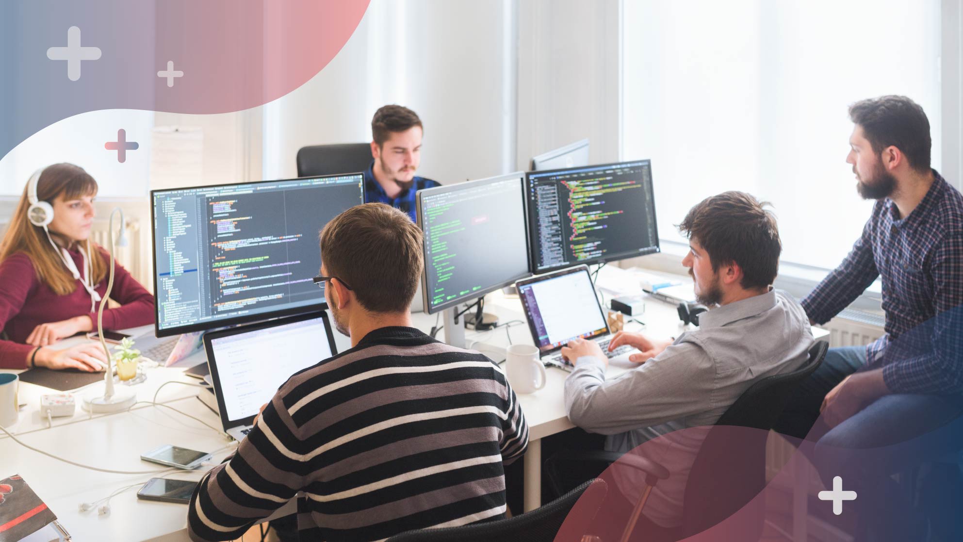 A group of five people work at a desk with multiple computer monitors. Two focus on code, one types on a laptop, one person listens with headphones, and another engages in discussion.