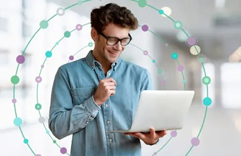 A man wearing glasses holds a laptop, smiling looking at the screen