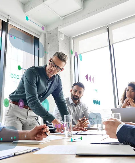 In a meeting room, a man points while others listen attentively around the table.