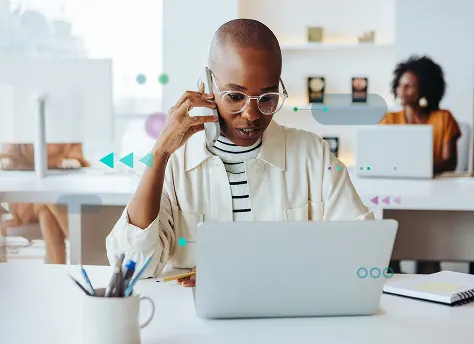 A woman sits at a desk with a laptop, engaged in a phone conversation.