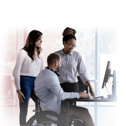 Three colleagues collaborating in an office. One man is in a wheelchair using a computer, while two women look on and discuss the work.