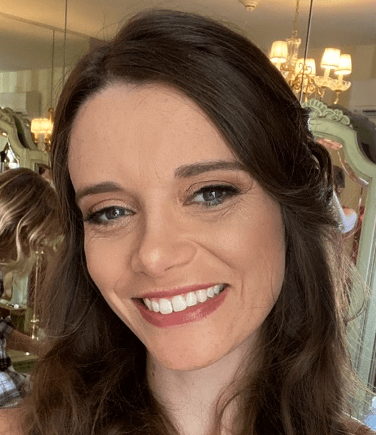 Headshot of blog author Chris Sheroff. She has long brown hair smiles at the camera in a room with ornate mirrors and chandeliers.