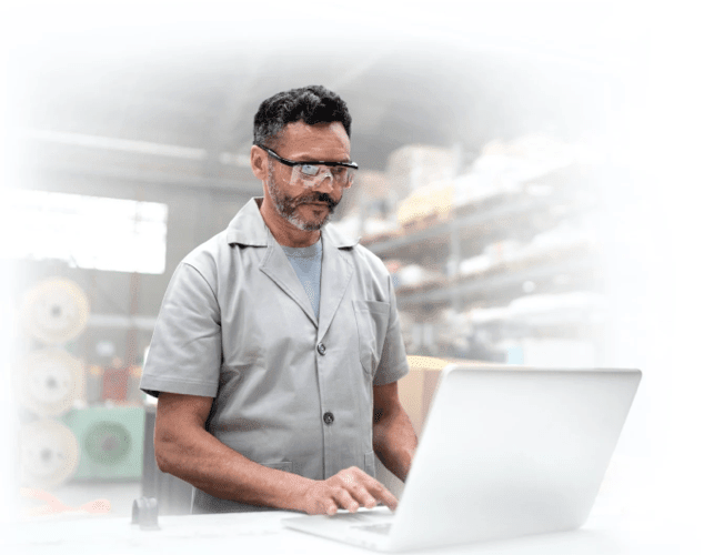 A man with safety glasses and a grey work shirt stands at a workbench using a laptop in an industrial setting. Shelves with materials are visible in the background.