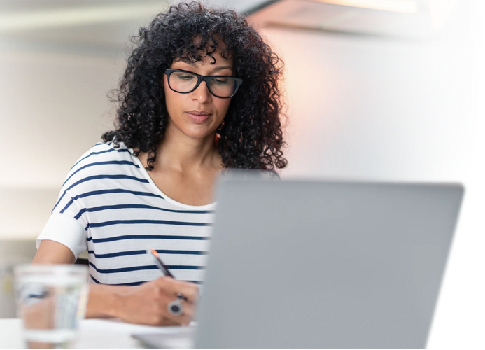 A person with curly hair and glasses is sitting at a desk, writing in a notebook while looking at a laptop. A glass of water is on the desk.