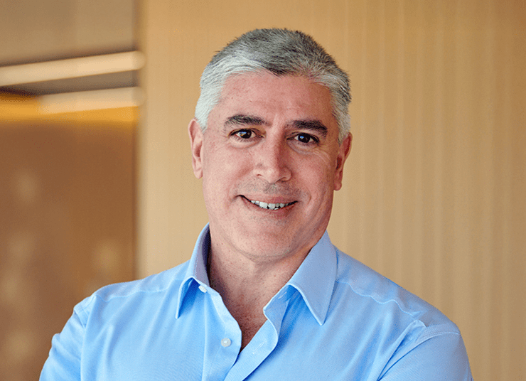 A headshot of a man with a warm, friendly smile, wearing a blue buttoned collar shirt. The background is softly blurred with warm, light brown tones.