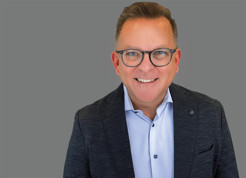 Matt Hayes, General Manager, Data Business Unit at Qlik and is wearing a dark textured blazer over a light-colored collared shirt stands against a plain gray background.