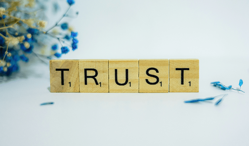 Scrabble tiles spelling "TRUST" are arranged on a white surface, with blue and white dried flowers partially visible in the background.