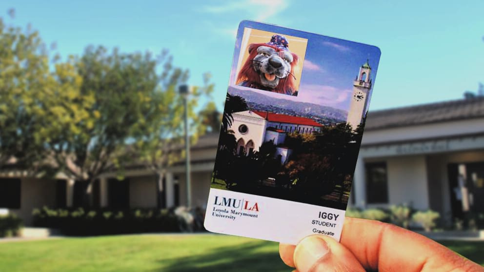 A hand holds a student ID card from Loyola Marymount University with a photo of a mascot lion named Iggy. The background features campus buildings and a clock tower.
